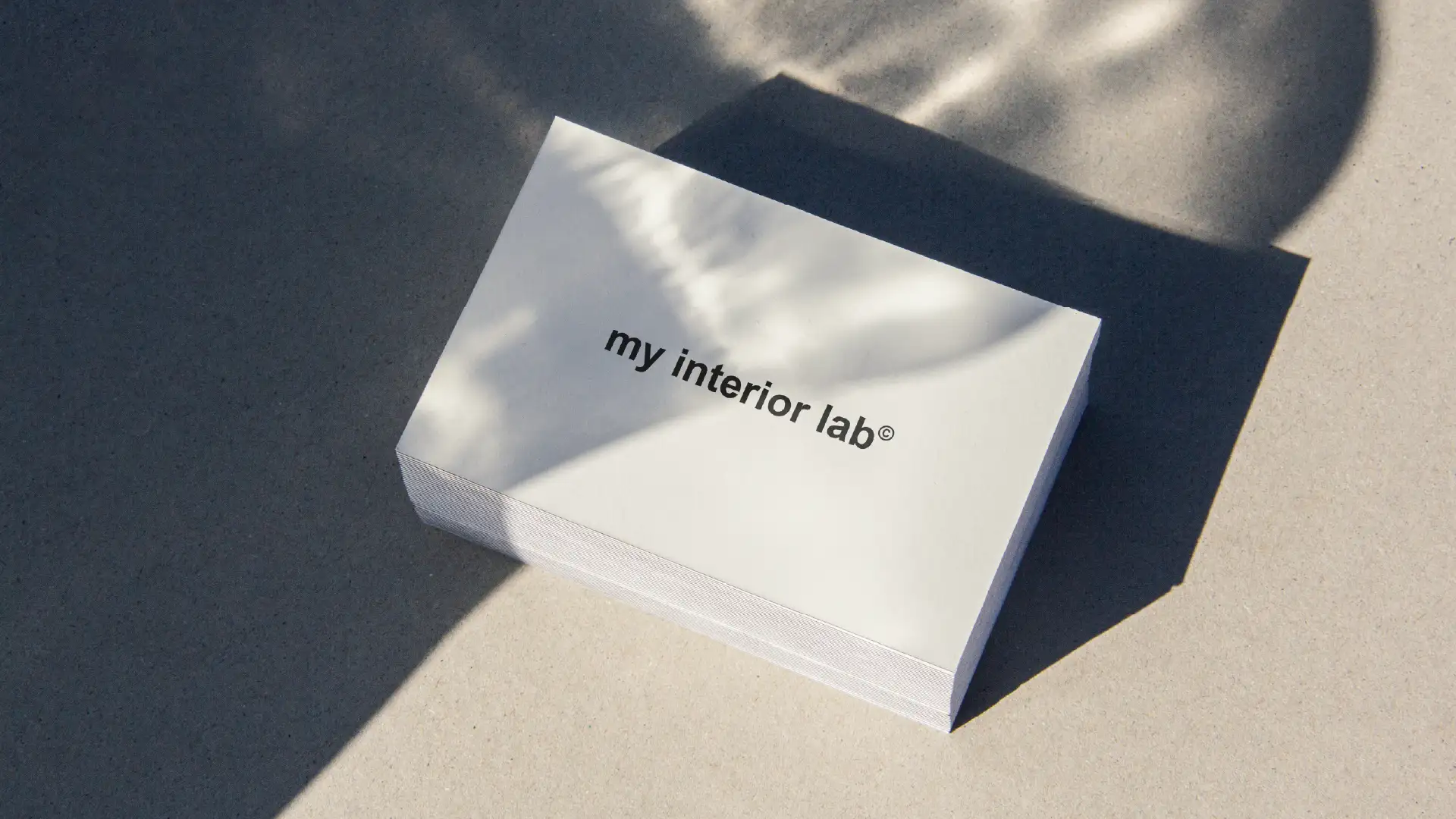 Close-up stack of My Interior Lab business cards with centered logo on cream, placed on smooth gray concrete floor, shadow and light play, minimalist aesthetic
