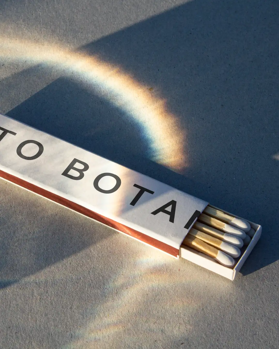 Close-up of elegant slim white Orto Botanico matchbox on raw concrete floor, light play creating subtle rainbow prism effect, refined minimalist design
