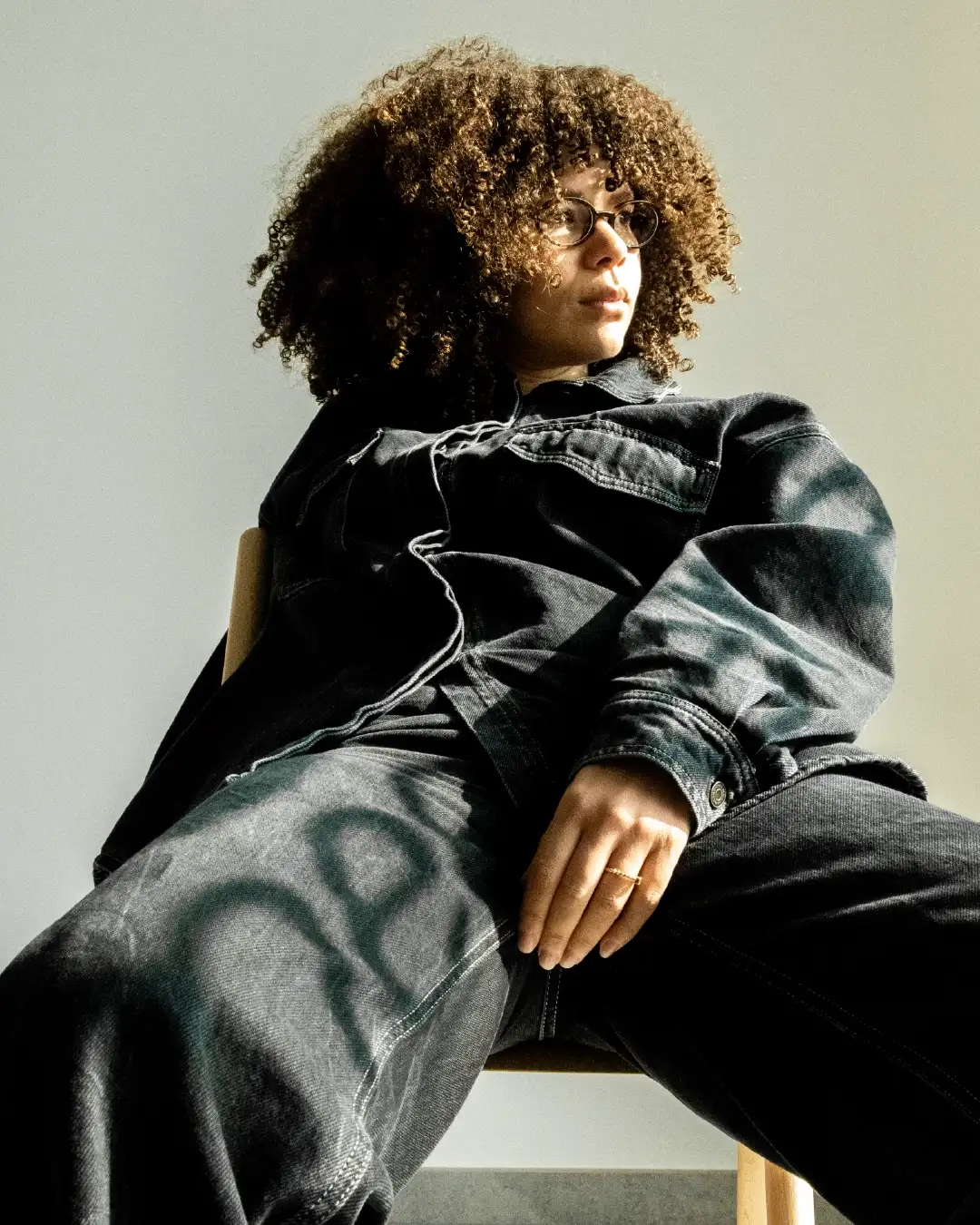 Portrait of Nééré founder Véronique Pirotton sitting casually looking aside in dark denim jumpsuit, Nééré logo reflected by light on garment, powerful minimalist design