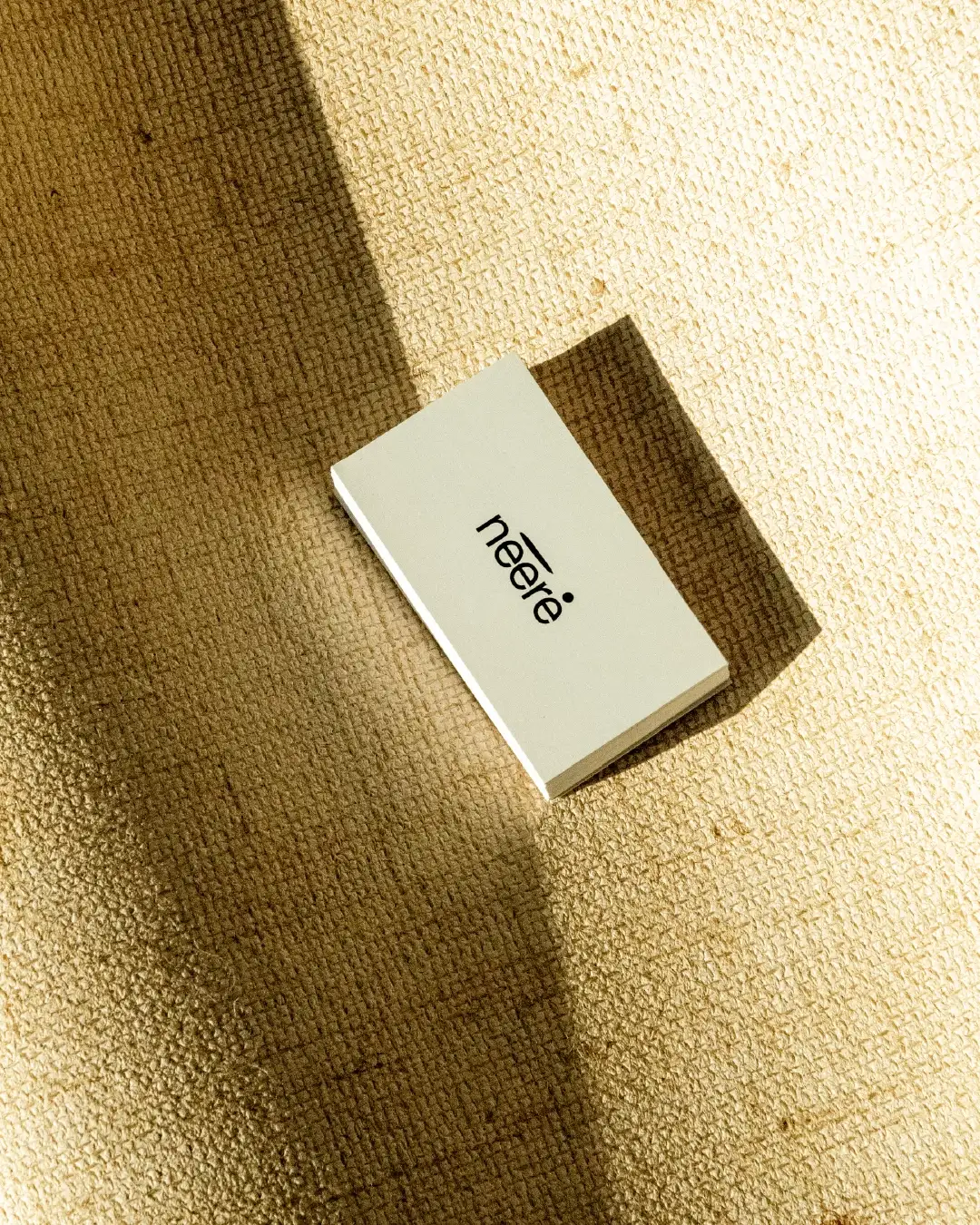 Close-up stack of Nééré business cards with centered logo on cream, placed on thick natural fiber textile, shadow and light play, minimalist organic atmosphere