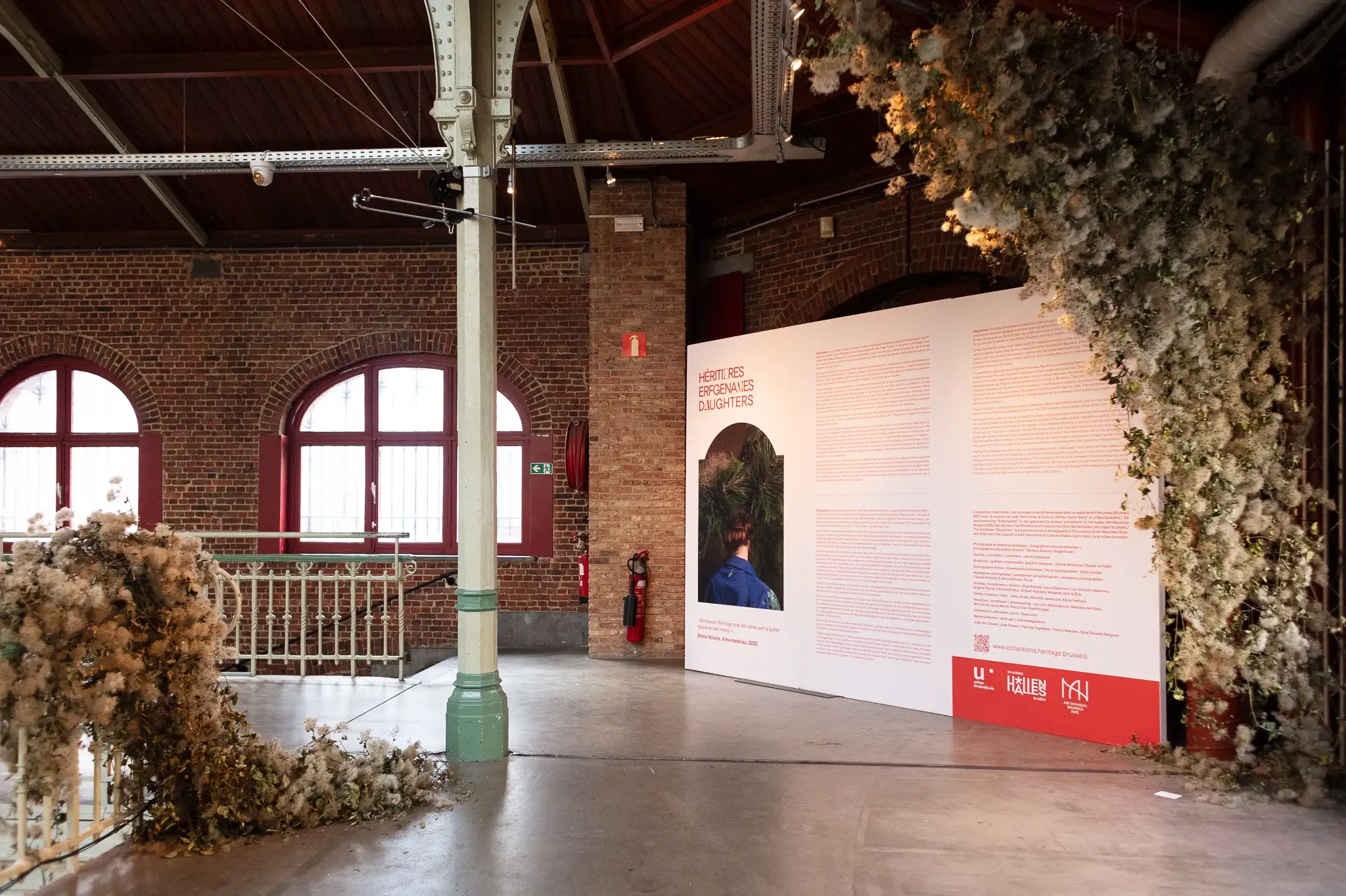 Héritières exhibition installation at Halles Saint-Géry Brussels by Barbara Salomé Felgenhauer: large white panel with red text blocks, master exhibition photo and partner logos, monumental floral compositions left side and ceiling