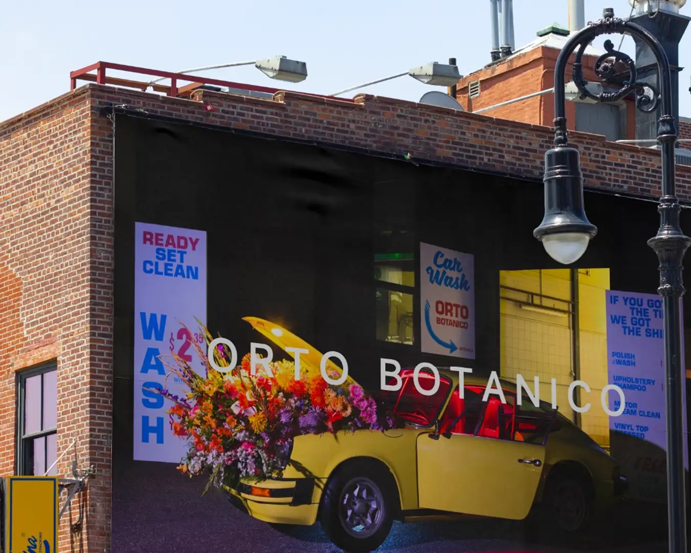 Billboard géant à Brooklyn sur mur de briques pour la marque Orto Botanico, montrant une scène de carwash années 70 avec une Porsche jaune ouverte remplie de fleurs colorées ; branding Studio La Folie.