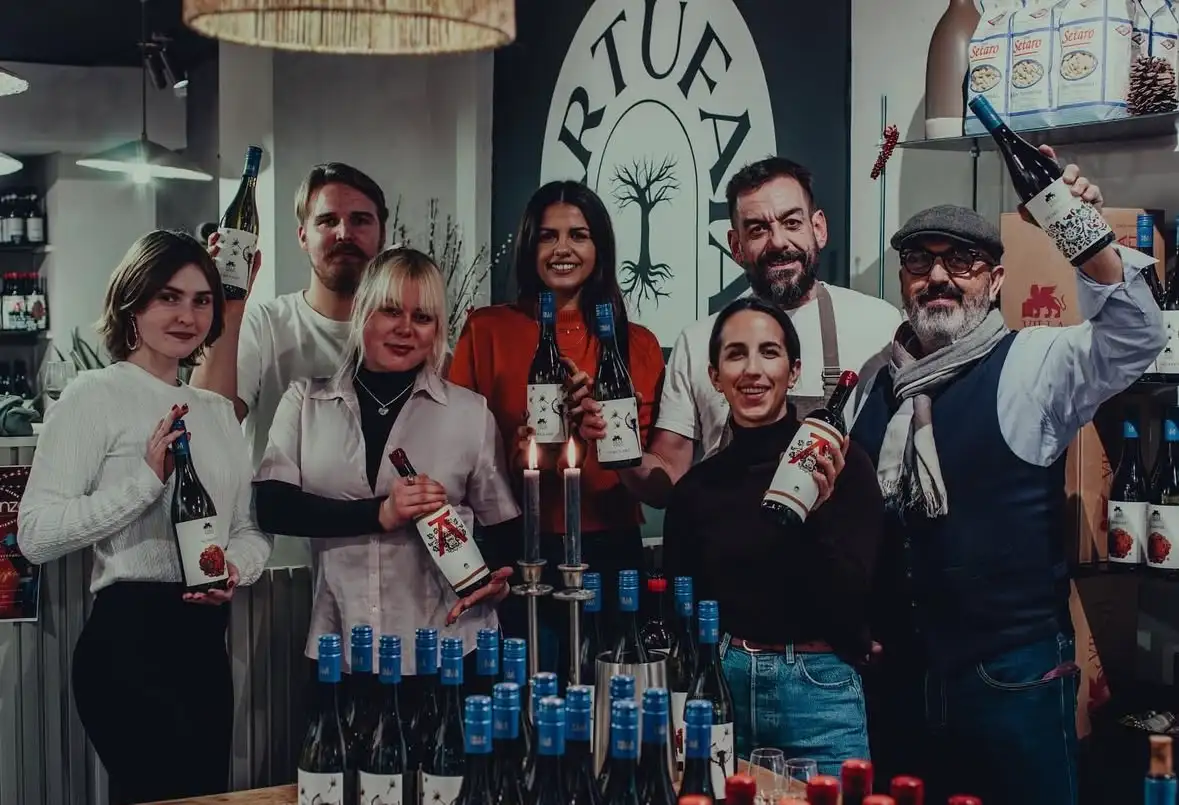 Équipe Tartufaia Maastricht sept personnes levant joyeusement bouteille de vin dans boutique, mur arrière avec logo crème peint à la main, ambiance conviviale authentique