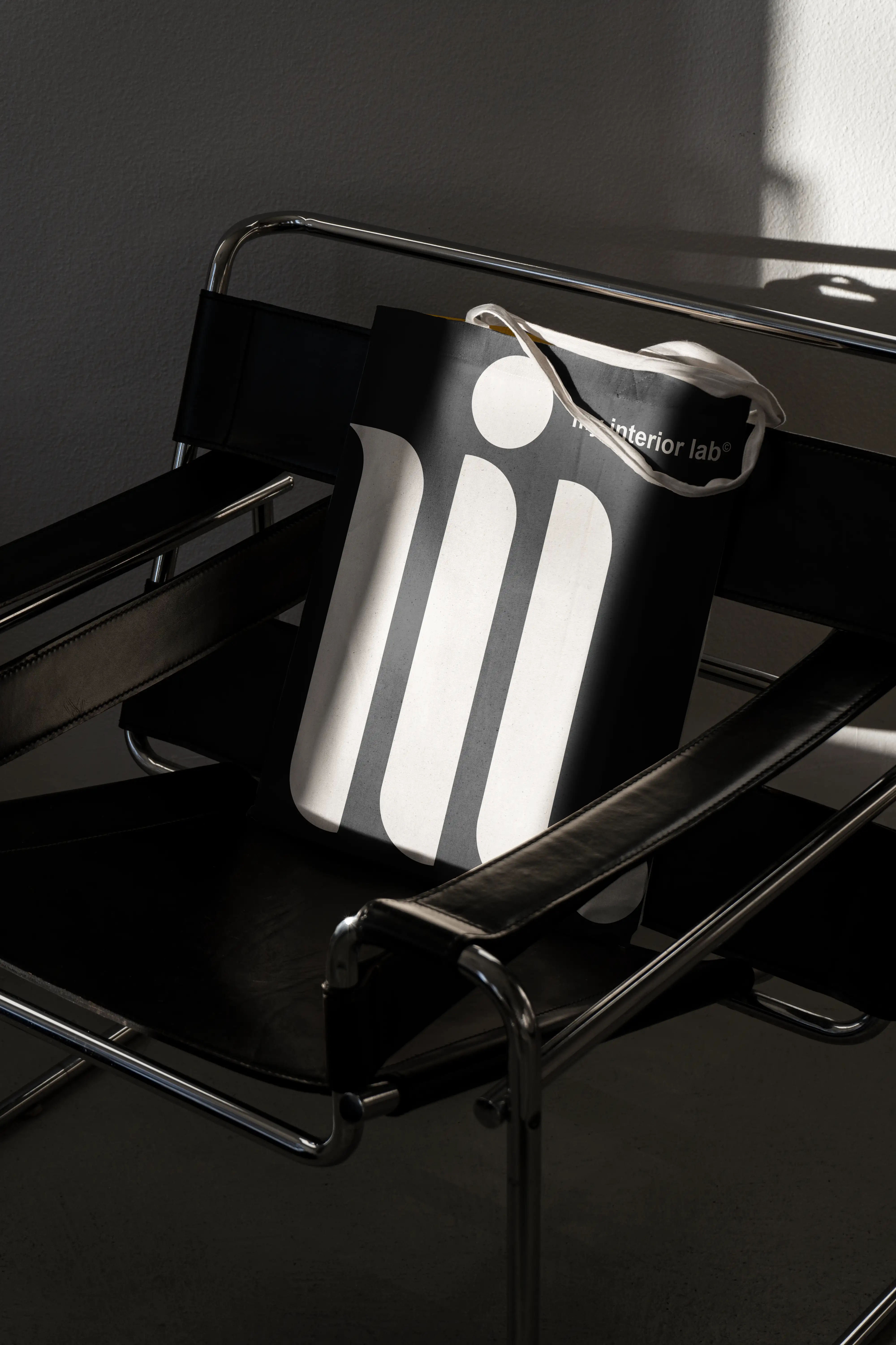 Close-up of black tote bag with oversized cream My Interior Lab monogram placed on black leather Le Corbusier-style armchair, refined design aesthetic