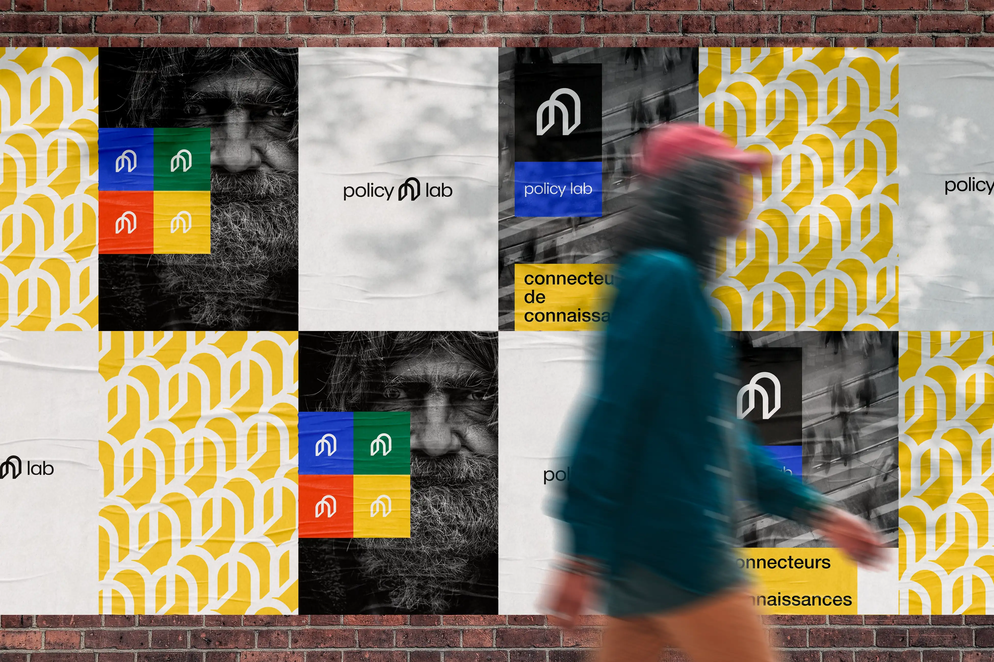 Twelve Policy Lab posters on red brick wall in two columns: white background logos, black-white bearded elderly man portraits with colored logo overlay, geometric brand pattern, walking pedestrian adding dynamic movement
